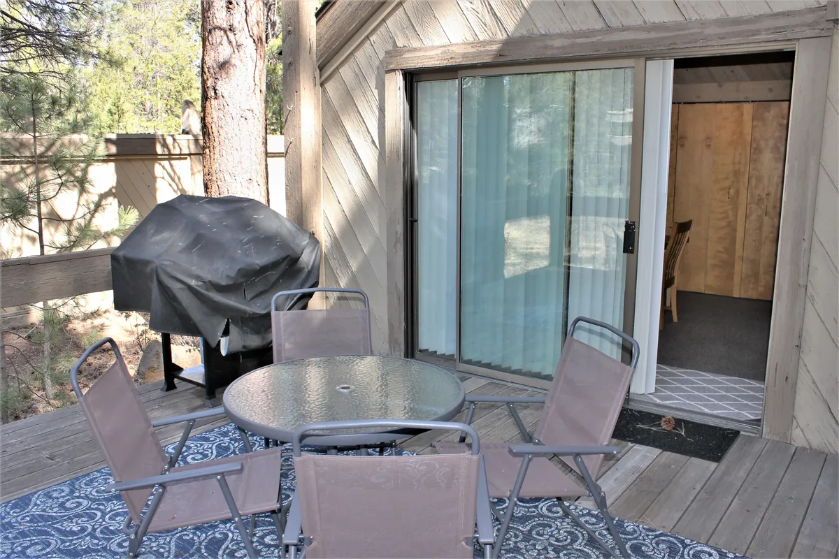 An exterior deck scene with a glass table, outdoor chairs, grill, and a sliding door that connects directly to the living space. The deck offers views of surrounding nature and provides an inviting spot for outdoor dining or enjoying coffee in the fresh air.