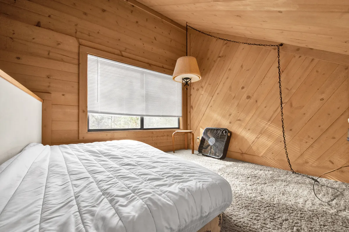 A serene bedroom with a large window lets in natural light and likely offers a view of the outdoors. The bed, lamp, and fan create a restful atmosphere, while the window acts as a focal point to the outside world.