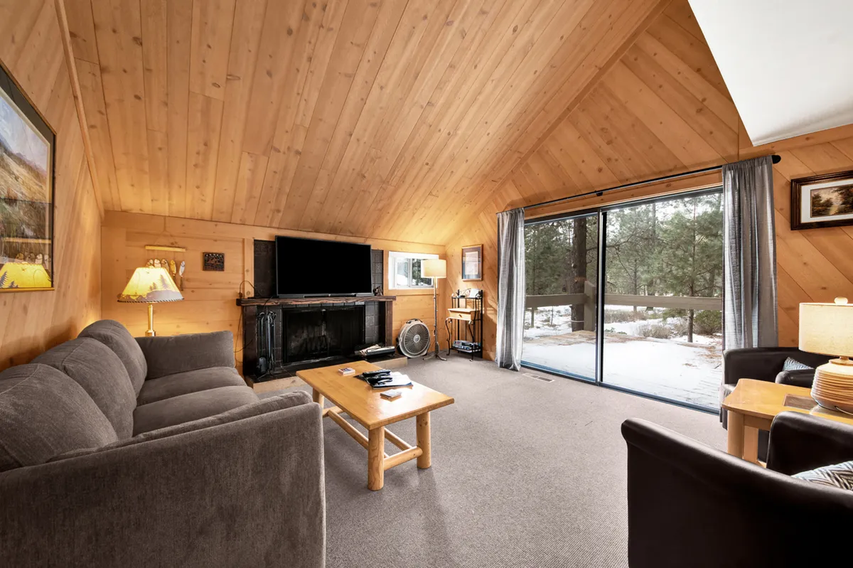 The image showcases a cozy living area with a plush sofa facing a glowing fireplace, large windows framing the outdoors, and a warm, inviting ambiance perfect for relaxing. The scene suggests a setting ideal for curling up with a book, sipping a warm drink, or simply unwinding while watching the snowfall outside.