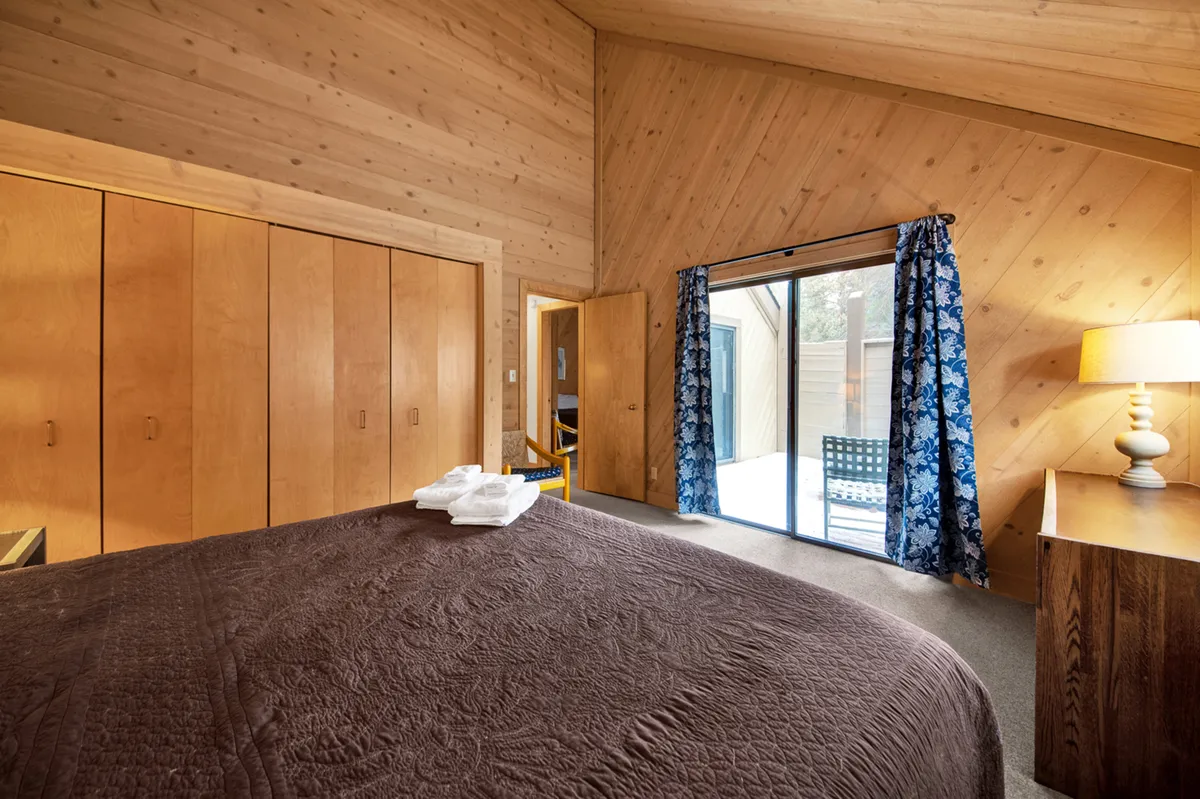 A serene bedroom boasting a large bed draped in a brown quilt, wooden storage cabinets, a sliding glass door leading to an outdoor patio, blue floral curtains, and a softly glowing bedside lamp. The layout suggests easy access and a peaceful setting for relaxation or morning routines.