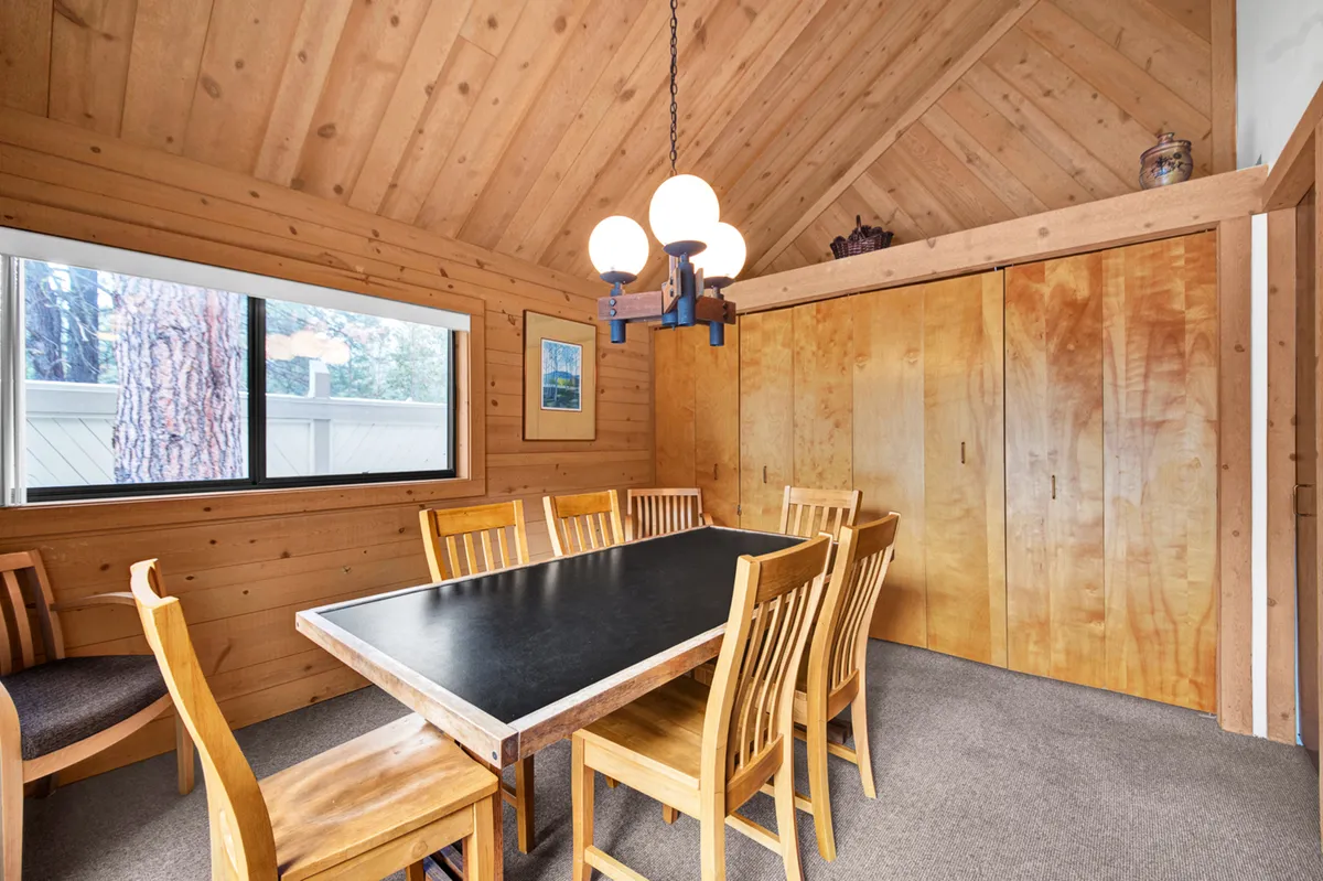 This image features a large dining table with wooden chairs placed near a window, offering a pleasant view outside. The chandelier overhead and the light streaming in suggest a cozy space that connects the indoors to the outdoors, perfect for enjoying meals while appreciating the scenery.