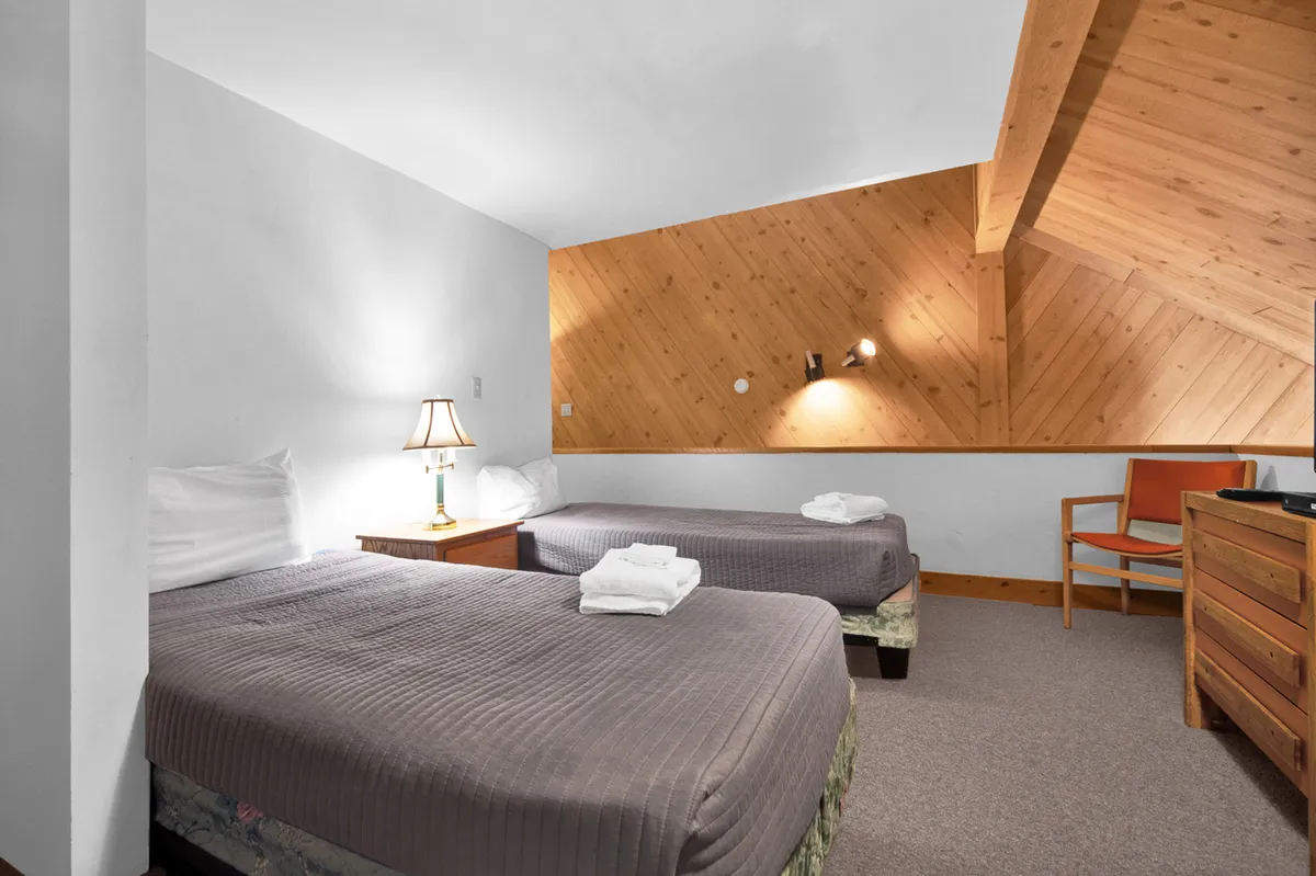 This image features two twin beds with a table lamp between them, nestled under a warm wooden ceiling. An armchair and a dresser add comfort and practical storage, with soft lighting creating a cozy, sleepover-ready atmosphere.