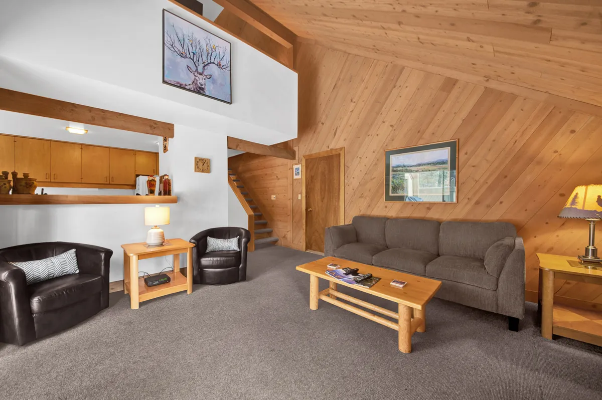 A cozy living room scene featuring a comfortable sofa, a wooden coffee table, exposed wooden beams, and artwork on the wall. Warm lighting and large windows create a welcoming, homey feel, perfect for relaxing, socializing, or enjoying a quiet moment by the fireplace.