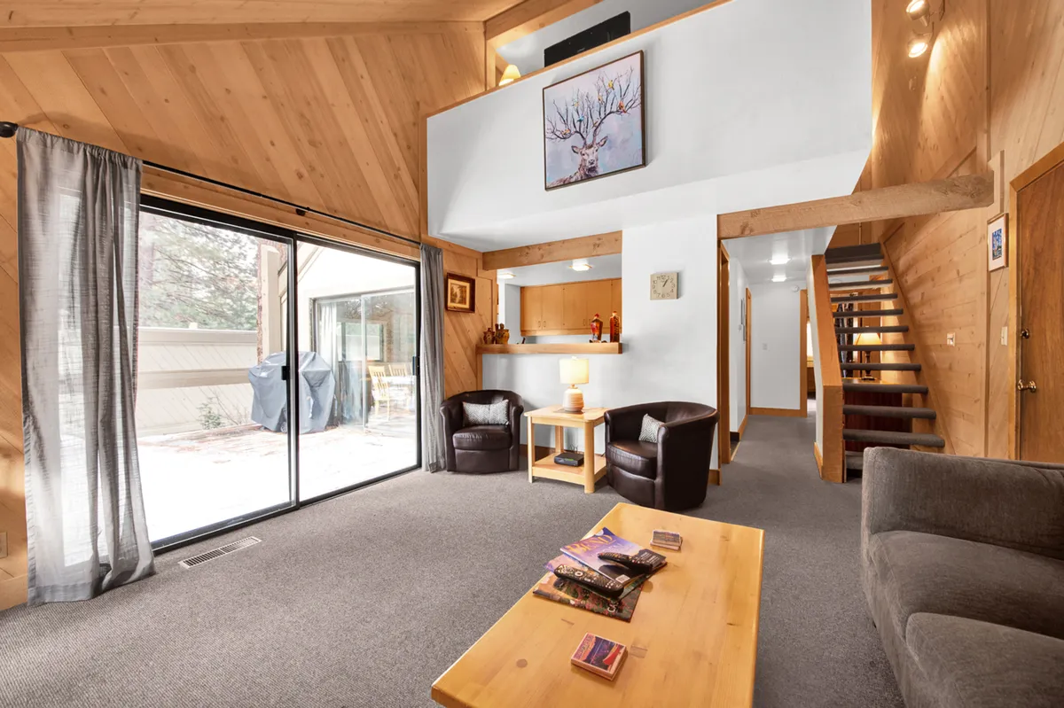 A bright living area featuring a large glass sliding door that offers a seamless view and access to the outdoors. Comfortable chairs and a wooden coffee table invite relaxation, while the staircase adds dimension and movement to the space. The natural light streaming in hints at the connection to trails and outdoor areas just beyond the door.